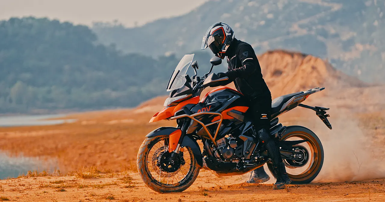Moto Zontes 350T X naranja y negra con piloto en casco y traje, en terreno de tierra con montañas y lago al fondo